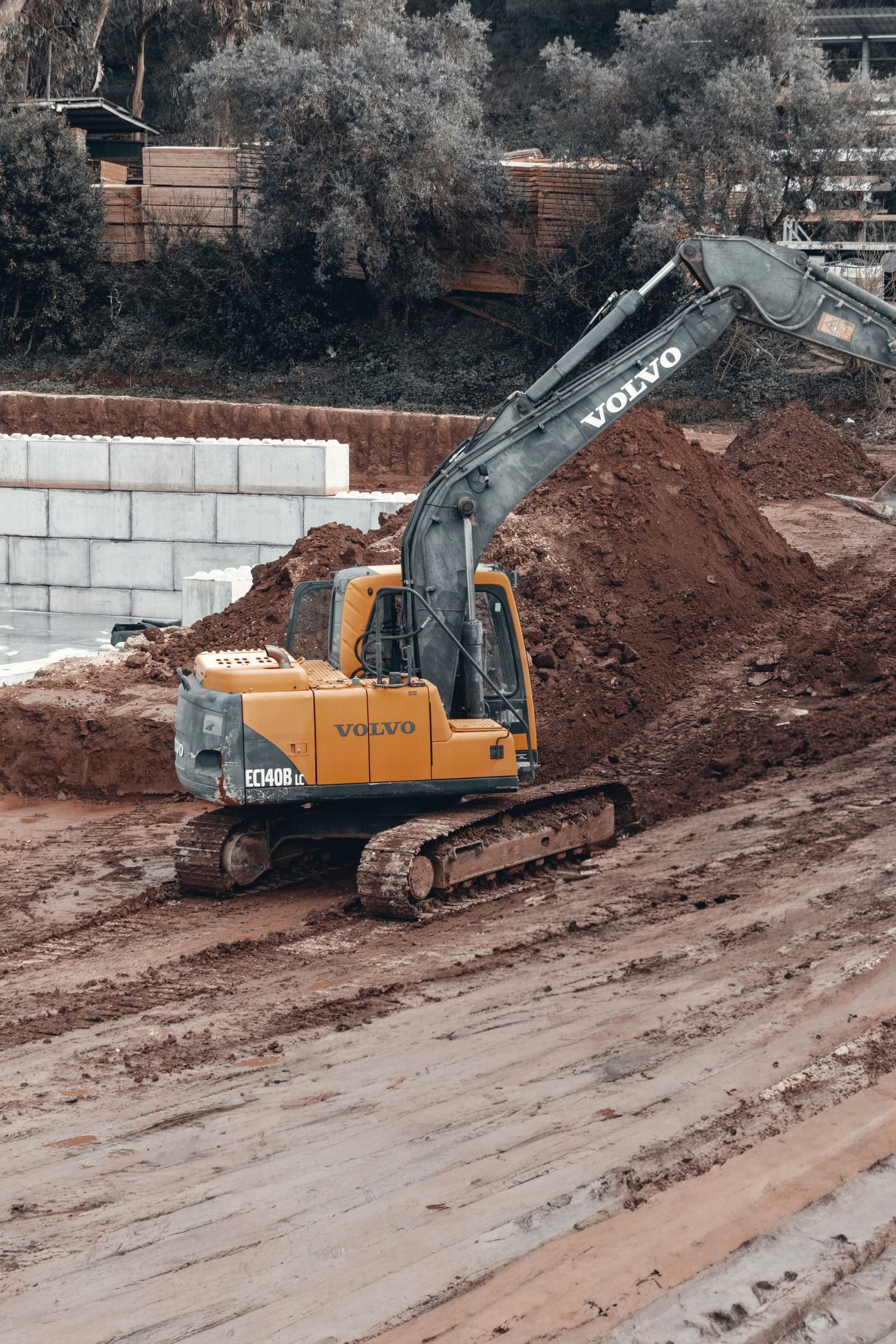 Excavator working a wet cut of red dirt beside a block wall